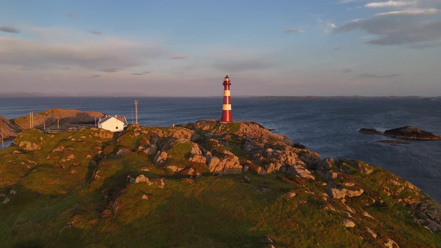 A beautiful red and white striped lighthouse sits on a rugged, grassy island overlooking the vast ocean during a golden sunset