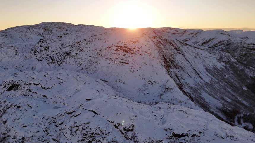 A majestic aerial view shows the sun rising over a vast, rugged mountain range completely covered in snow