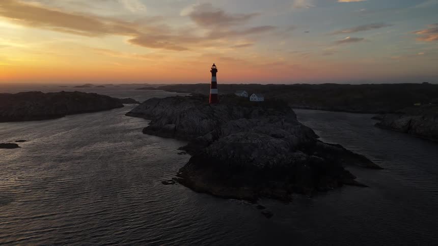 A serene aerial pullback reveals a remote lighthouse on a rocky coastline during a stunning sunset