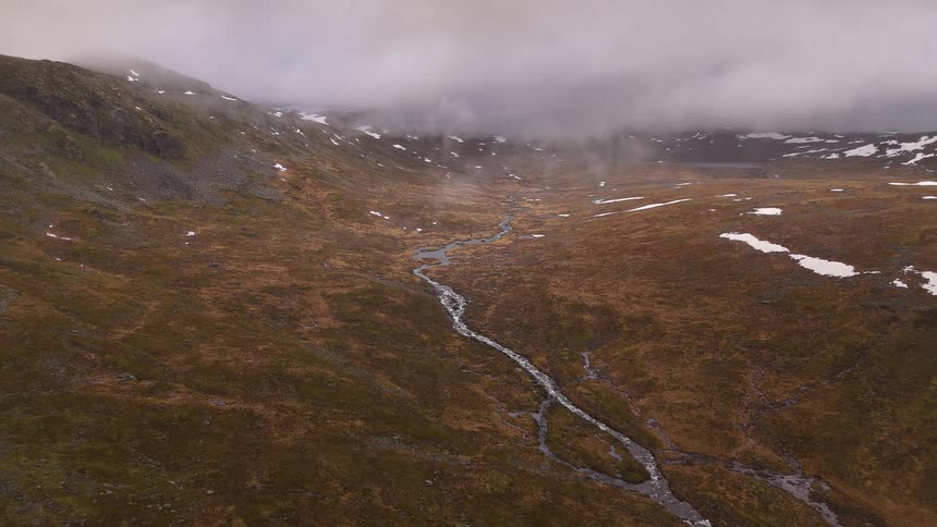 A static aerial view shows a winding stream flowing through a desolate, brown mountain valley with patches of snow under low, heavy clouds