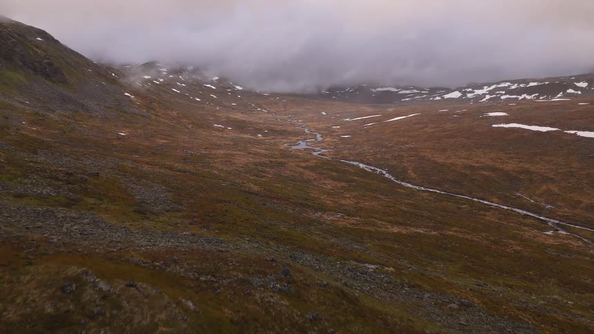 A slow aerial reveal of a stark and misty mountain valley with a winding stream and remnants of snow