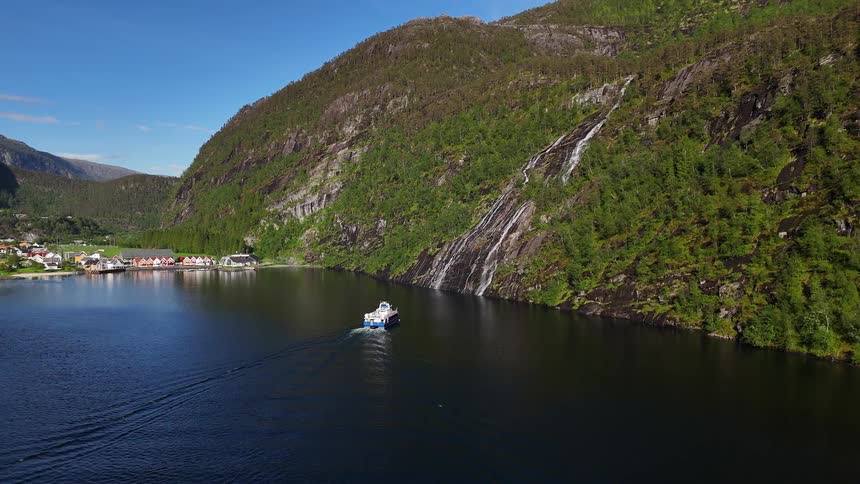An aerial view captures a ferry sailing through a stunning Norwegian fjord, with a waterfall on the lush mountainside and a quaint village on the shore