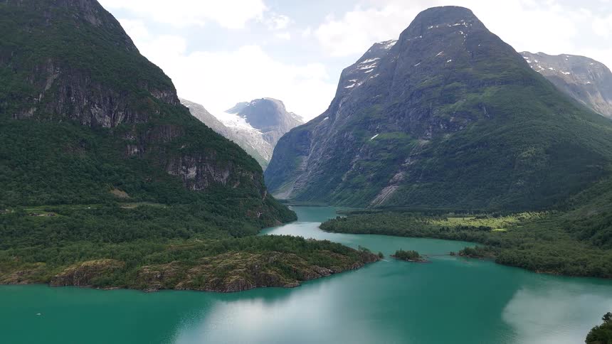 A breathtaking aerial shot reveals a vibrant turquoise glacial lake nestled within a valley of lush green mountains, with a massive glacier visible in the distance
