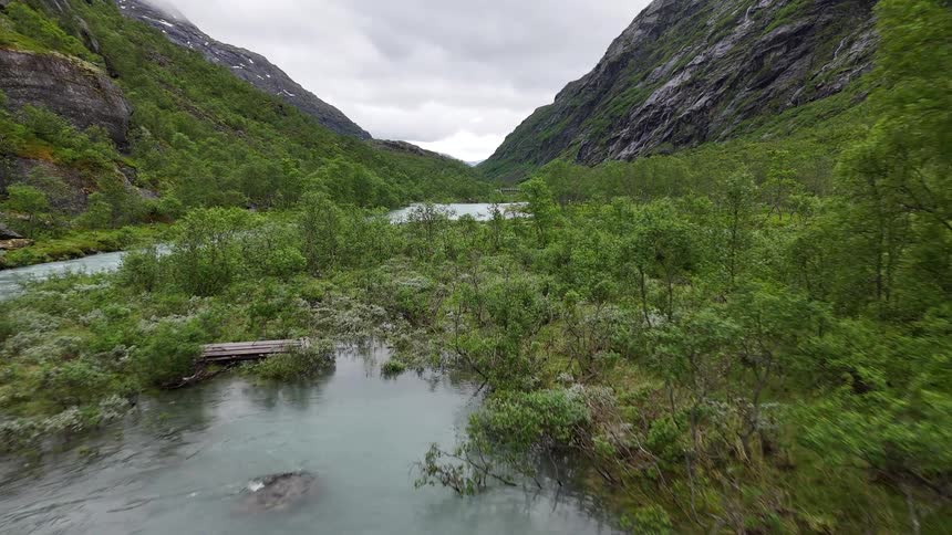 An aerial journey over a turquoise glacial river flowing through a lush, green valley surrounded by majestic mountains in Norway