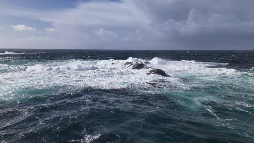 A dramatic slow-motion shot of powerful, foamy waves crashing over rocks in a turbulent ocean under a cloudy sky