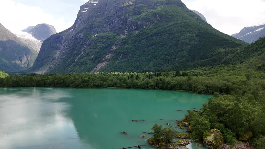 Majestic aerial pullback reveals kayakers exploring a brilliant turquoise lake at the base of a glacier-capped mountain