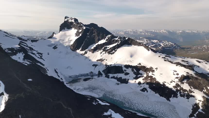 A static aerial view of a majestic snow-covered mountain peak with a glacier and a partially frozen turquoise alpine lake