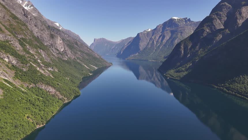 A serene, static aerial view captures the majestic beauty of a Norwegian fjord, where calm waters reflect the steep, green mountainsides and clear blue sky