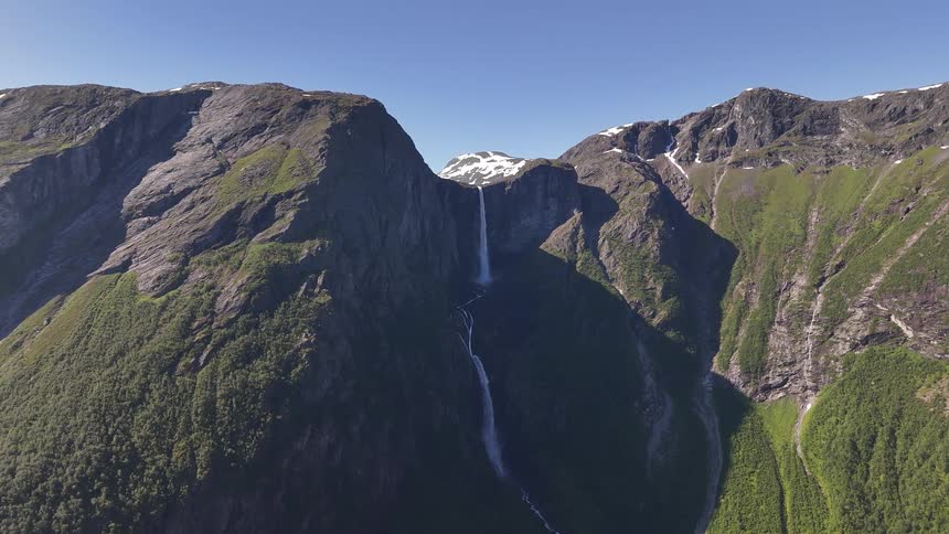 Static aerial view of a majestic waterfall cascading down a rugged mountain cliff into a lush green valley on a sunny day