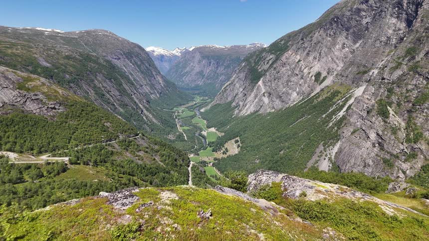An aerial view reveals a lush green valley with majestic mountains and snow-capped peaks in the distance