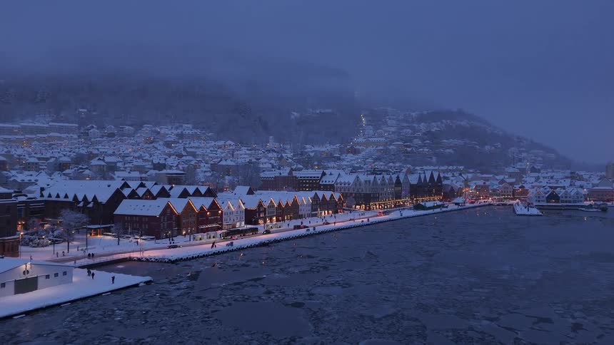 A serene winter evening in Bergen, Norway, with the snow-covered Bryggen wharf lit up beside a frozen harbor