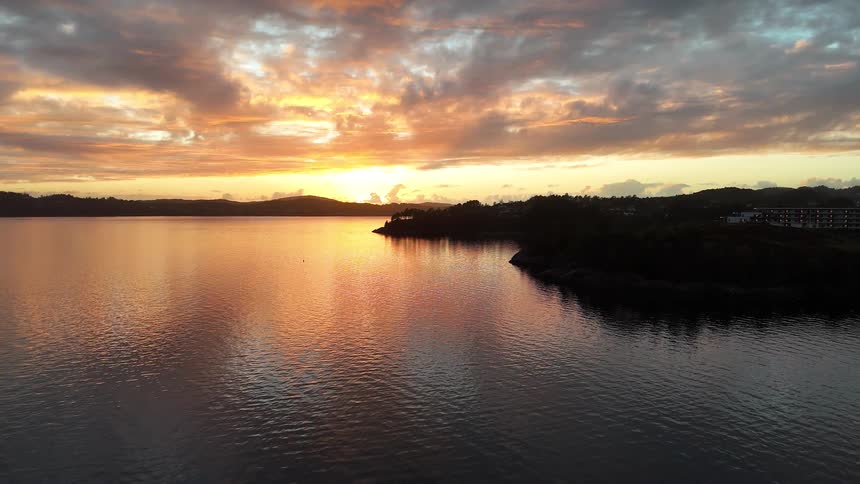 A tranquil aerial view shows a vibrant sunset reflecting on the calm water of a bay, with a silhouetted coastline in the distance