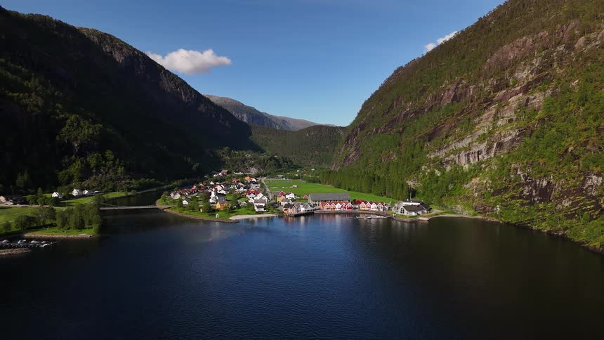 A beautiful Norwegian village with colorful houses sits peacefully on the shore of a dark blue fjord, framed by towering green mountains on a sunny day