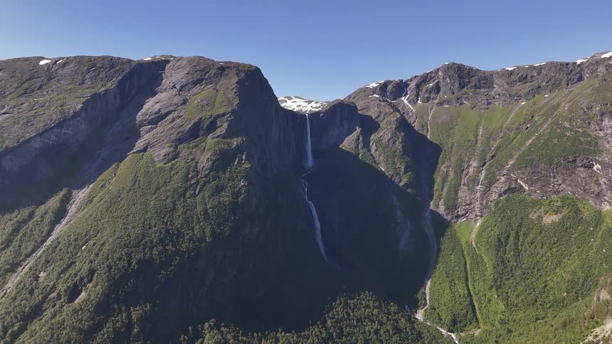 A stunning aerial view shows the majestic Mardalsfossen waterfall flowing from a snow-capped mountain peak down into a verdant, sunlit valley