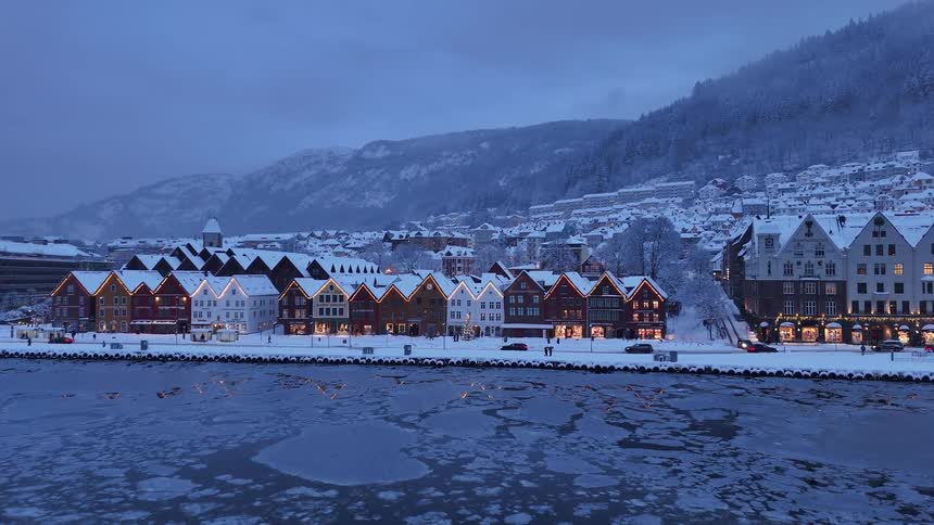 The iconic Bryggen wharf in Bergen, Norway, glows with festive Christmas lights during a snowy winter twilight, with ice floating on the calm harbor waters