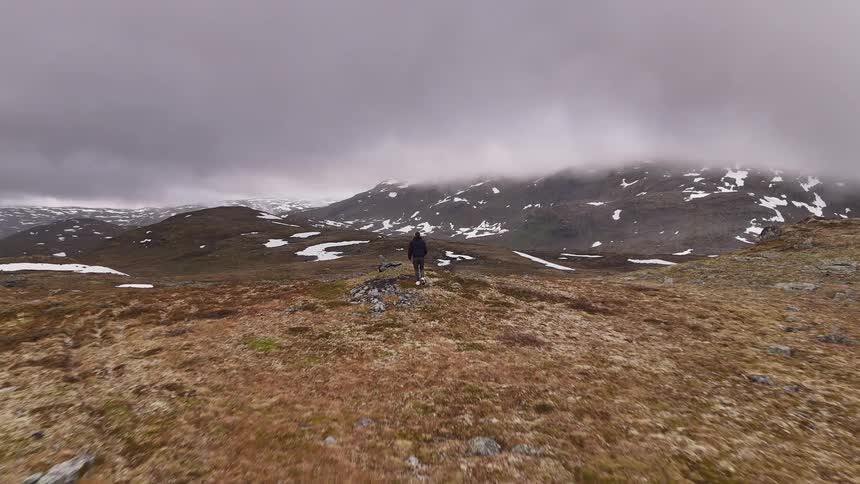 Following a lone hiker from behind as he walks through a vast, rocky landscape with snowy mountains under a gloomy sky