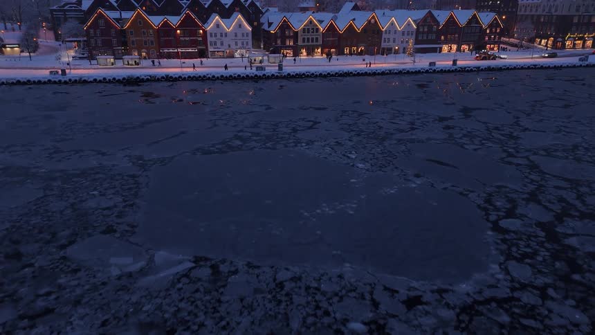 An aerial shot flies over a frozen harbor at twilight to reveal the beautifully illuminated and snow-covered historic buildings of Bryggen in Bergen, Norway