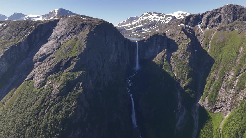 A wide aerial view of a majestic waterfall flowing from snow-capped mountains in a lush green valley on a clear sunny day