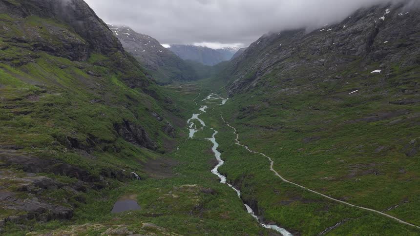 An epic aerial view pulls back over a vast green valley carved by a winding glacial river, nestled between towering, cloud-covered mountains