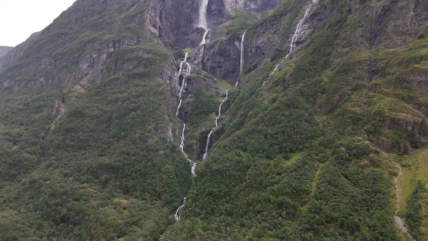 A stunning aerial view tilts up to reveal multiple waterfalls cascading down a steep, verdant mountainside in Norway