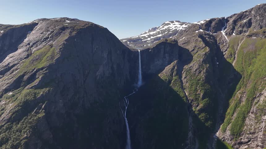 A majestic static aerial view of the breathtaking Mardalsfossen waterfall cascading down a rugged, green mountain valley with snow-capped peaks in the distance