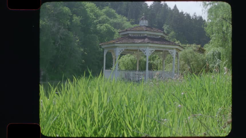 A vintage 16mm film pan reveals a charming white gazebo by a tranquil pond surrounded by lush greenery