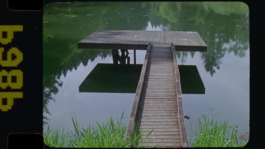 A nostalgic vintage 16mm film shot slowly tilts up to reveal a serene wooden dock on a calm lake with two people sitting underneath