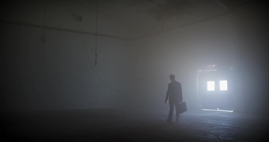 A silhouetted man in a suit holding a briefcase emerges from a bright doorway into a dark and foggy warehouse