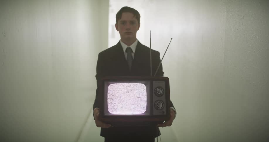A young man in a suit mysteriously appears in a sterile white hallway, holding an old television with a static screen