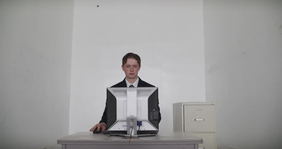A young businessman works at an old computer in a stark, white, empty office before staring blankly at the camera