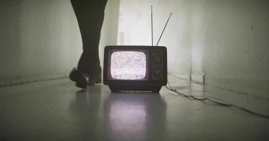 A mysterious man walks down a long hallway and stops behind an old TV with a static screen