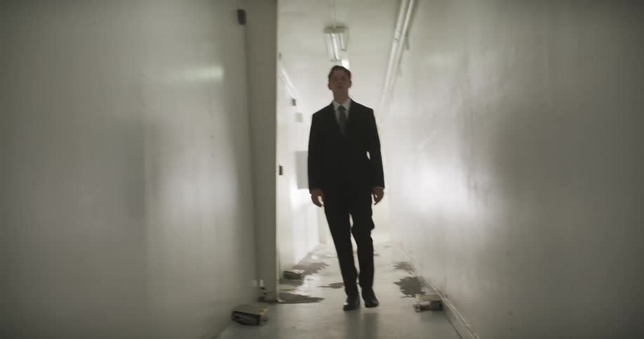 A man in a black suit walks in slow motion towards the camera down a long, derelict hallway as the light fades to darkness