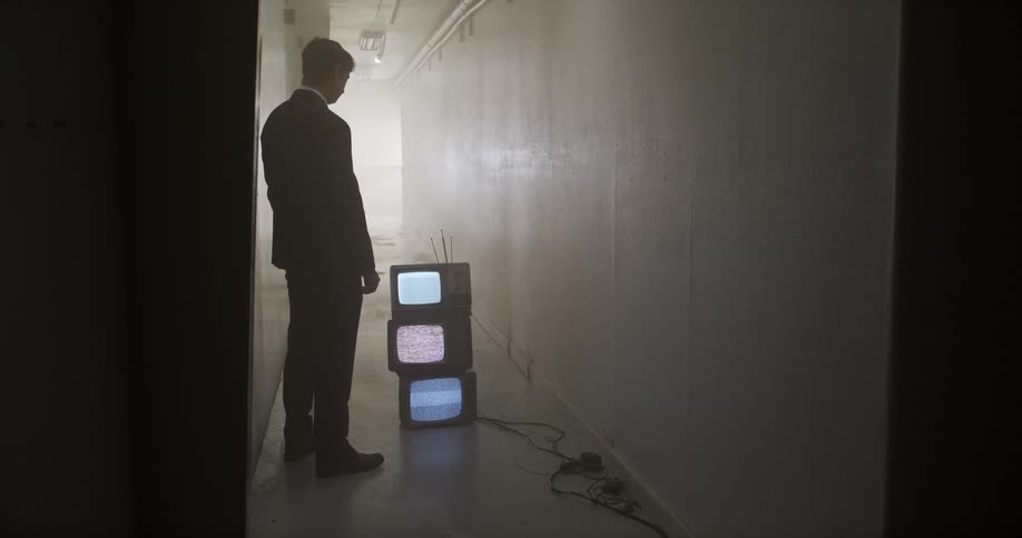 A silhouetted man in a suit watches a stack of three retro TVs displaying static in a dark, atmospheric hallway