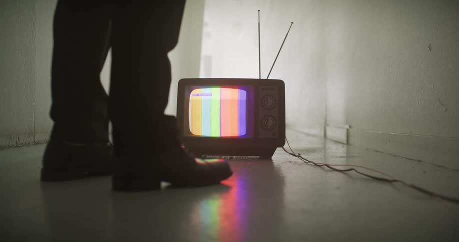 The feet of a person are silhouetted in a dark hallway by the glow of an old television displaying a colorful 'no signal' test pattern