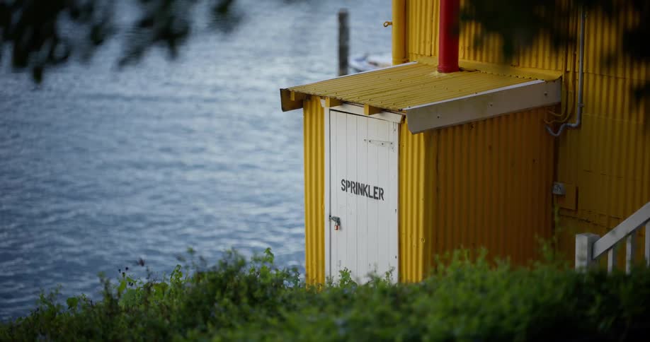 A yellow corrugated metal shed with a 'Sprinkler' room door sits next to a calm body of water