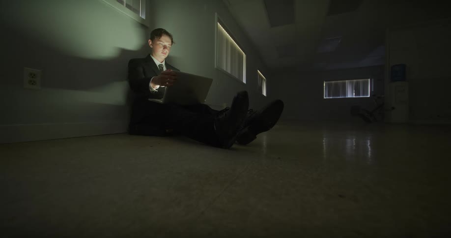 A young businessman in a suit works late at night, sitting on the floor of a dark and empty office while using his laptop and phone