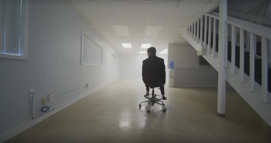 A businessman in an empty, sterile office rights an overturned chair, spins in it, and then playfully rides it across the floor