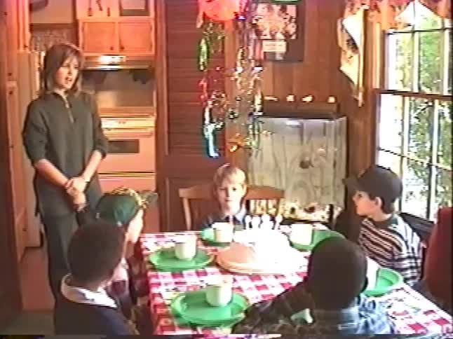 A young boy sits with his friends at the dining room table in front of a birthday cake with lit candles