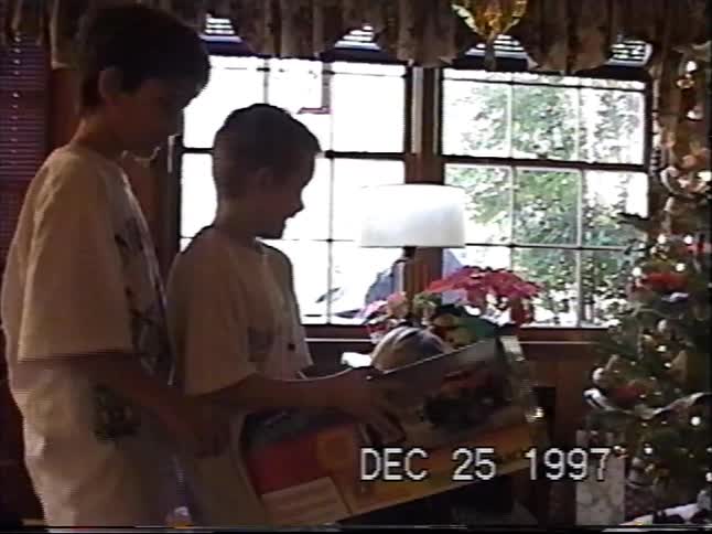 A young boy excitedly opens a large toy for Christmas next to his brother in a 1997 home video