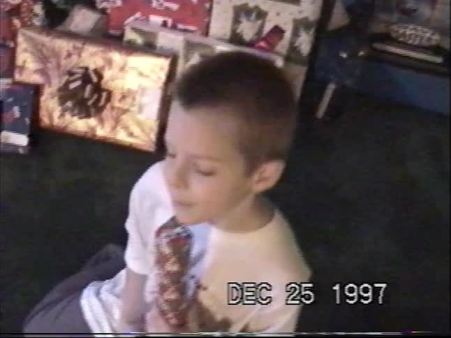 Young boy sits on the floor opening a wrapped Christmas present beside a tree and stacked gifts in a 1997 home video