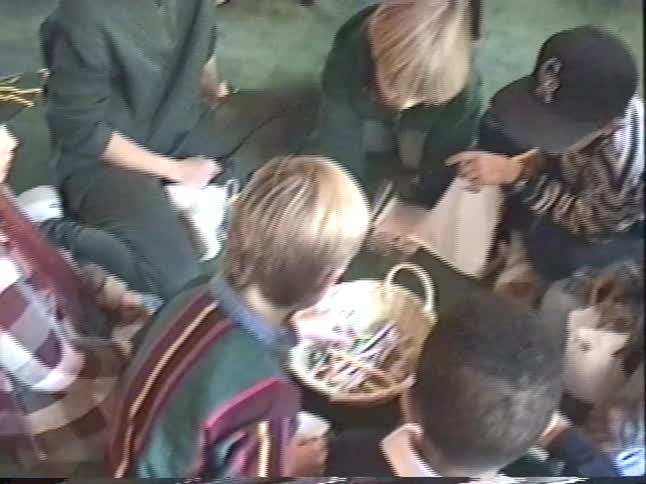 A vintage home video pans from women talking on a couch to children sitting on the floor looking through their Easter eggs and baskets