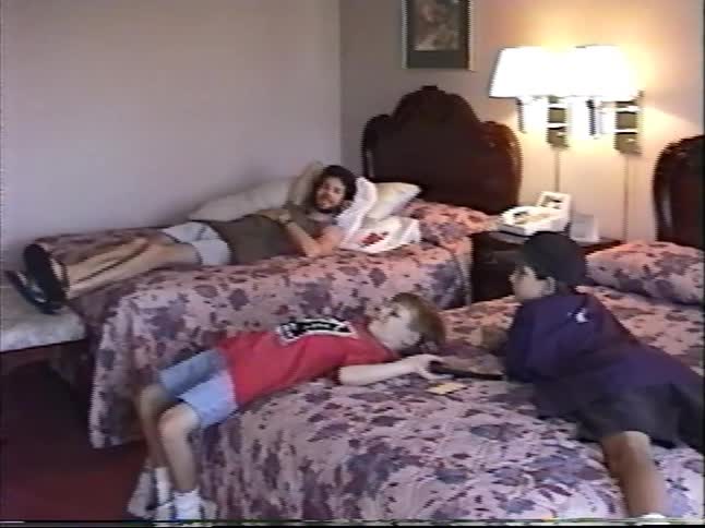 Vintage VHS footage shows a father and his two children relaxing on beds in their hotel room during a family vacation
