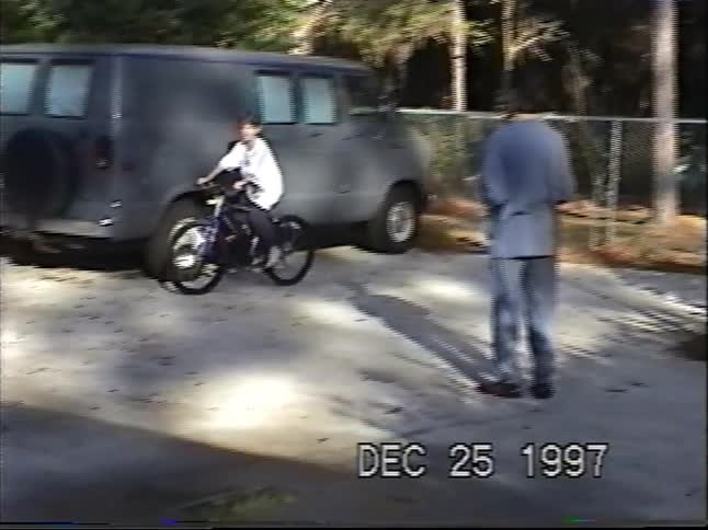 A vintage home video captures a young boy riding his brand new bicycle in the driveway on Christmas morning in 1997