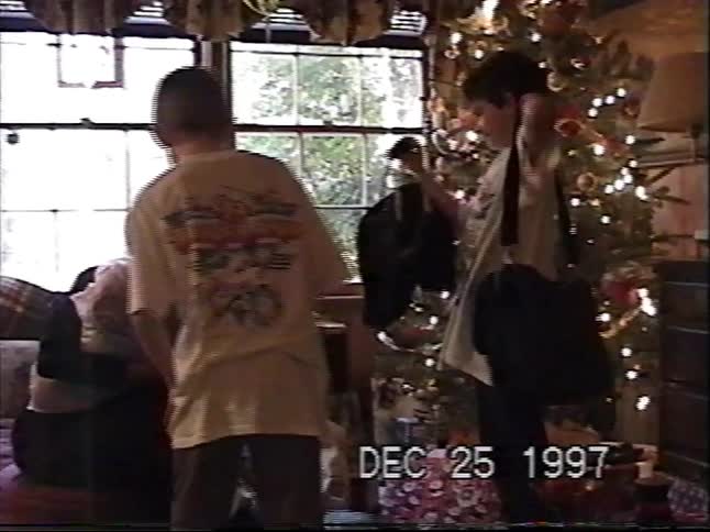 A young boy excitedly opens presents by the Christmas tree with his family in a nostalgic home video from 1997