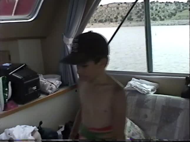 Boy in a baseball cap and towel stands inside a boat cabin with lake views through the windows