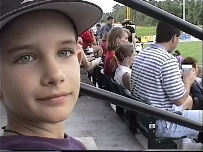 A playful young boy with bright eyes hides behind his baseball glove while watching a game from the stadium stands