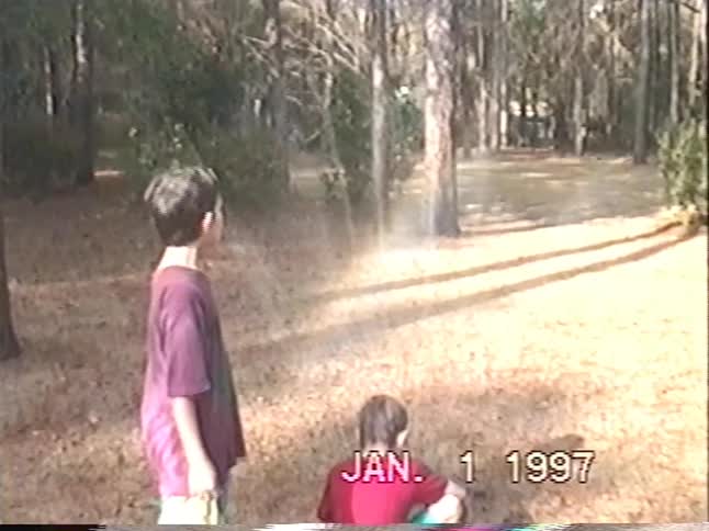 Vintage 1997 footage shows two young boys having fun playing in a water sprinkler in a wooded backyard, creating a small rainbow in the mist