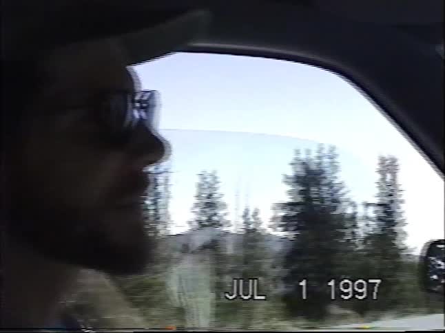 A vintage VHS recording shows a family driving on a winding mountain road through a forest on a sunny day in 1997