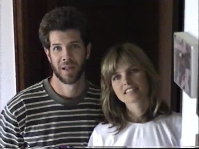 A young couple walks down a hallway and poses for the camera, eventually breaking into laughter in a classic 1990s home video