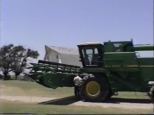 A farmer performs maintenance under his large green combine harvester on a sunny day at a rural farm